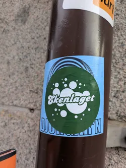 This is a sticker on a pole. The sticker has a light blue background, and a green circle with white dots and text. The text reads 'Ekenlaget DJURGÅRD'N'.