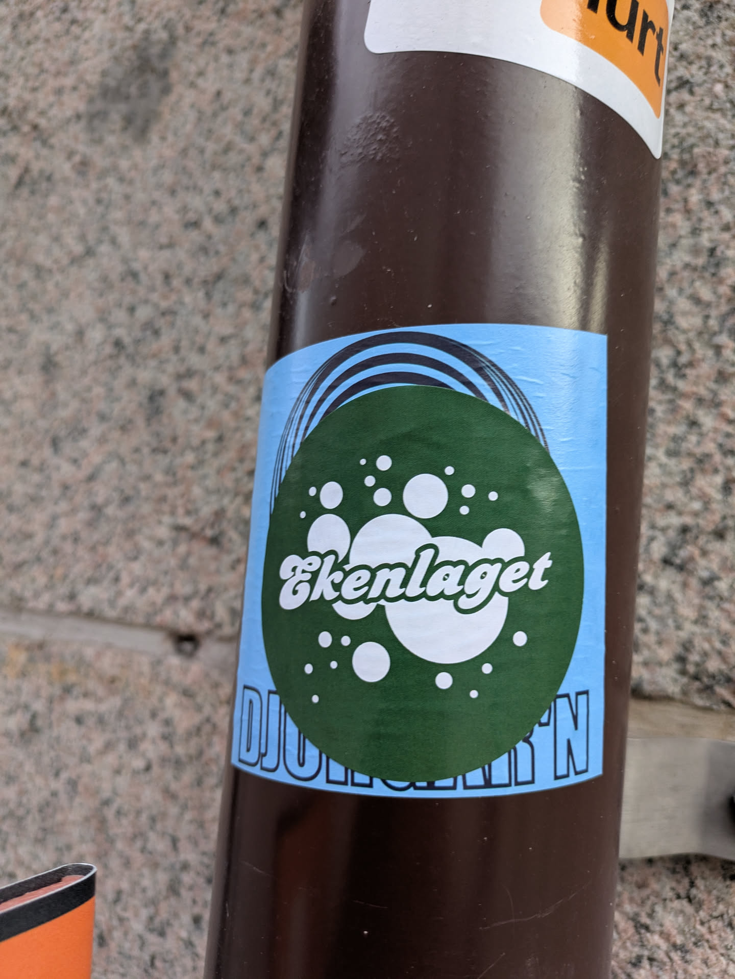 This is a sticker on a pole. The sticker has a light blue background, and a green circle with white dots and text. The text reads 'Ekenlaget DJURGÅRD'N'.