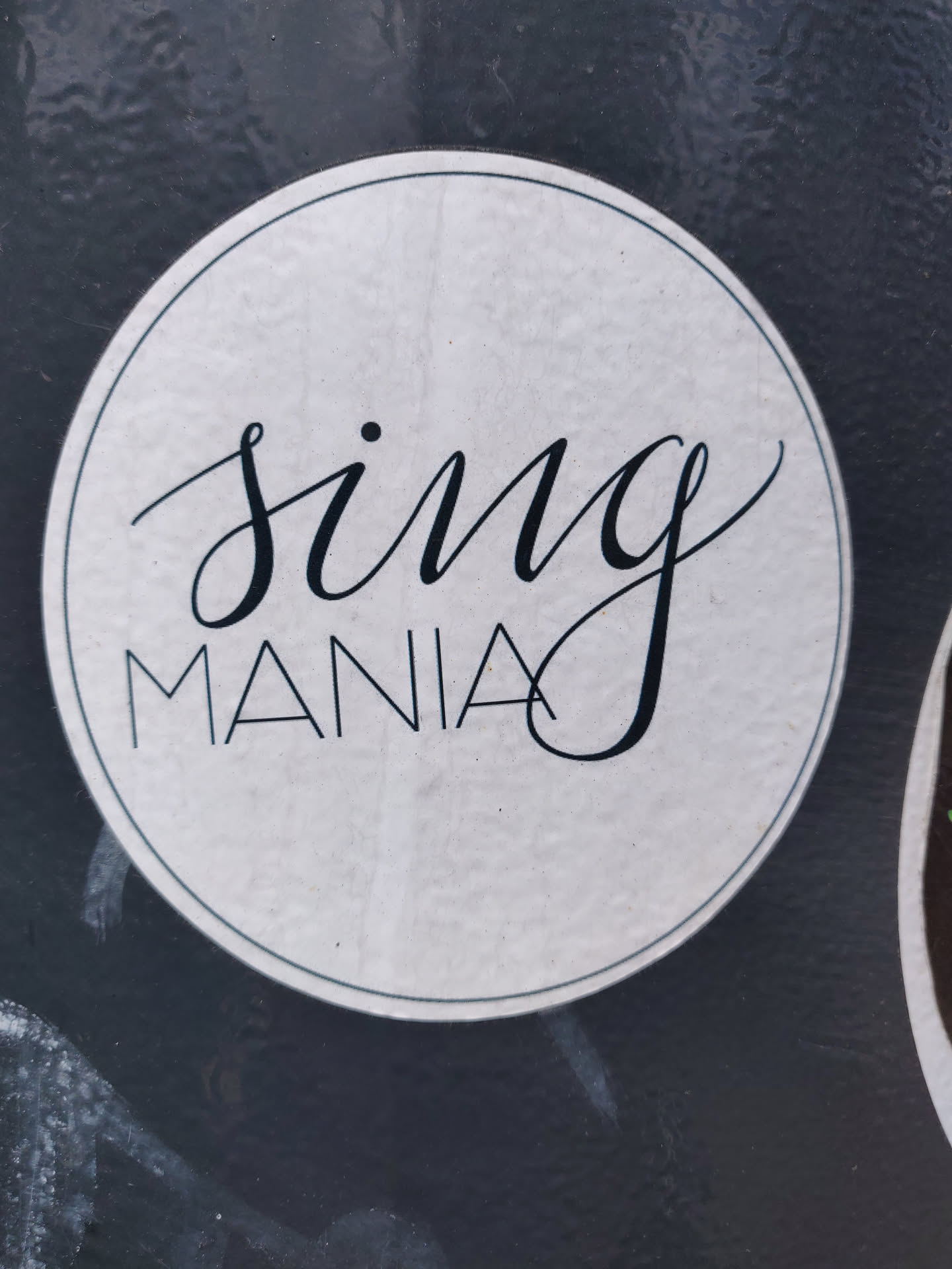 A round sticker with the text "Sing Mania" written in an elegant, stylized font. The text is in black on a white, textured background, which is circled by a thin black border.
