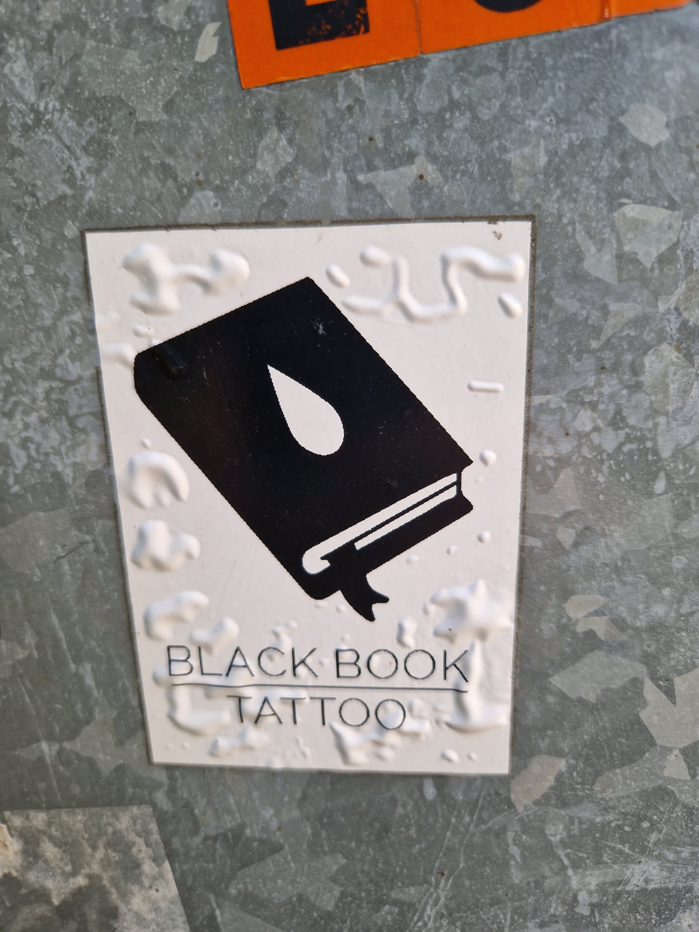 A sticker featuring a minimalist design of a book with a single teardrop. The text 'BLACK BOOK TATTOO' is written beneath the illustration.
