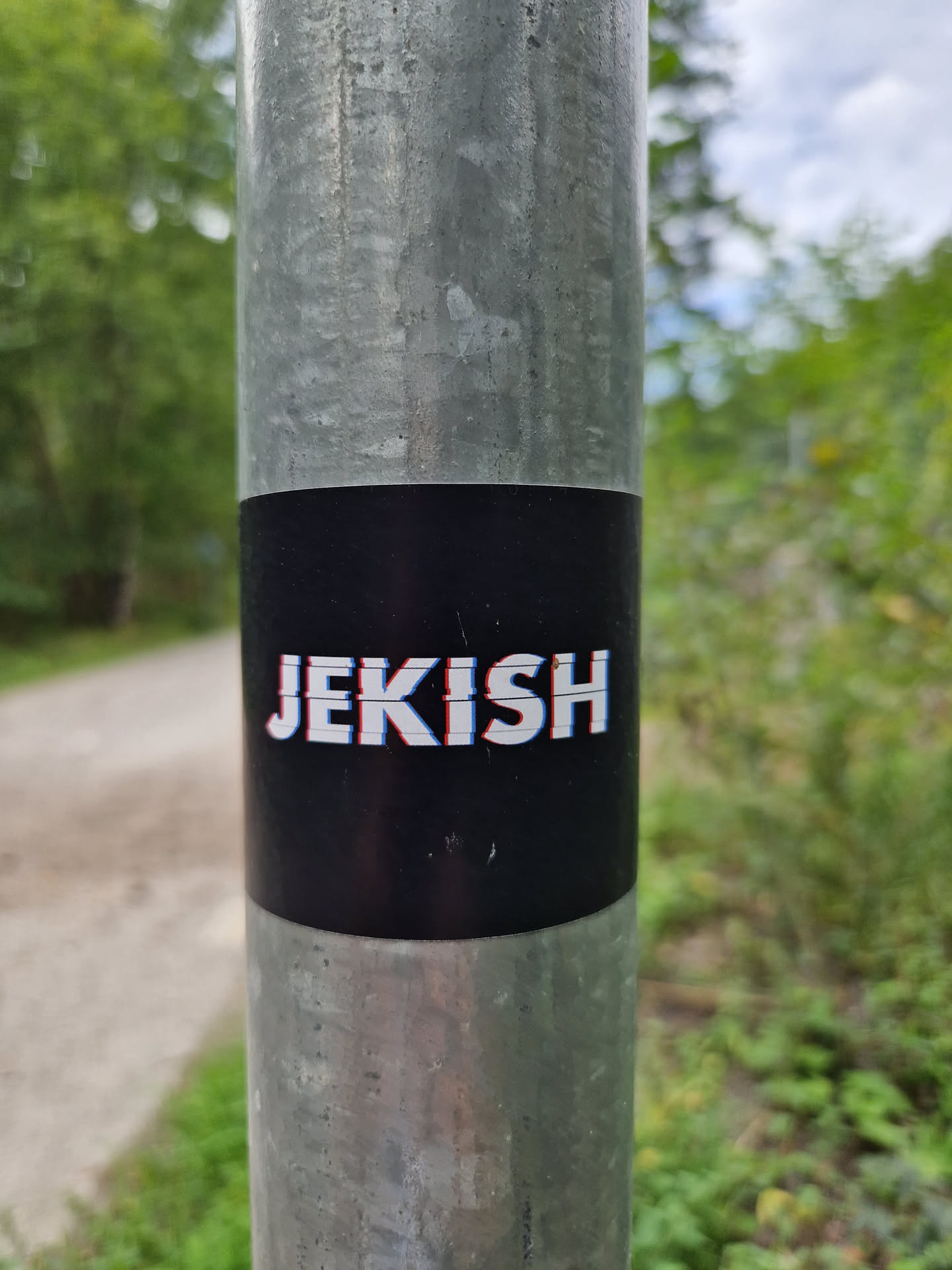 A black sticker with the word Jekish in a slightly glitched, white and light blue font. It's adhered to a metal pole outdoors.