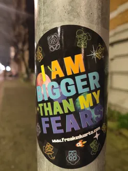 A round sticker with a black background and colorful text. The text says "I AM BIGGER THAN MY FEARS". There are also several small illustrations around the text. A website address is visible at the bottom: www.freakzduartz.com