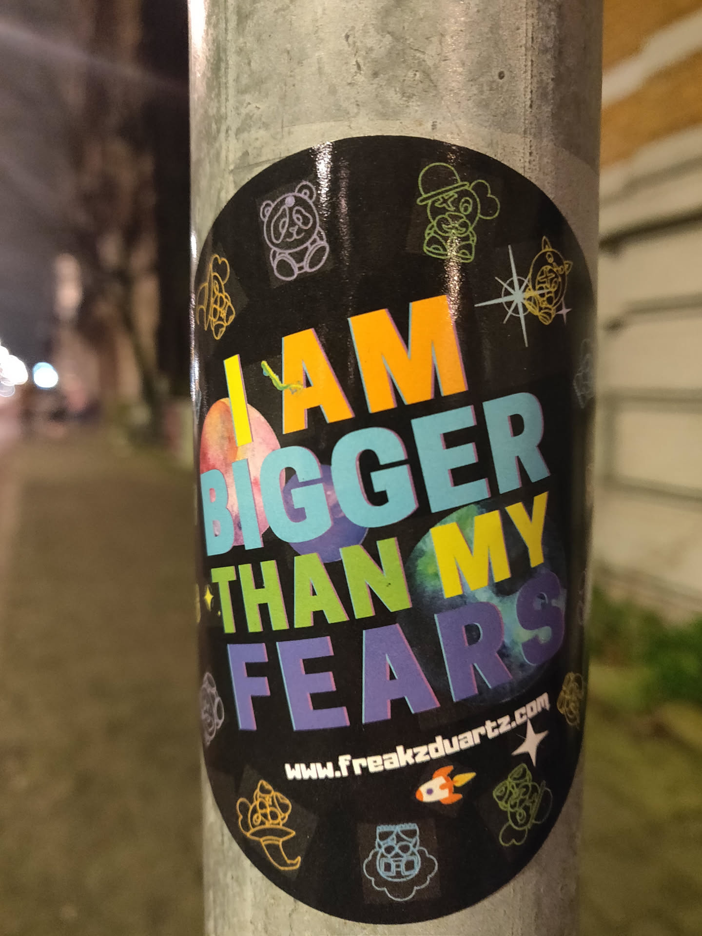 A round sticker with a black background and colorful text. The text says "I AM BIGGER THAN MY FEARS". There are also several small illustrations around the text. A website address is visible at the bottom: www.freakzduartz.com