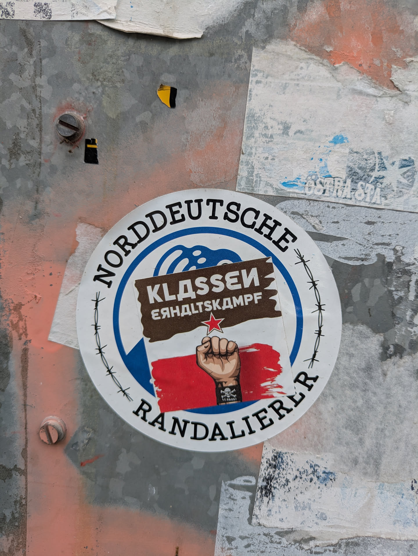 A circular sticker prominently displayed on a textured metal surface. The sticker has a white outer border with black text 'NORDDEUTSCHE RANDALIERER' (North German Rioters/Hooligans) encircling the design. Inside, a blue background with a stylized wave pattern at the top frames a central image. This image consists of a brown rectangular banner with white text 'KLASSENKAMPF' (Class Struggle) and a red five-pointed star. Below the banner, a raised, clenched fist with a black wristband featuring a white skull and crossbones (Jolly Roger, associated with FC St. Pauli) is depicted over a red, brushstroke-like background. A black barbed wire design encircles the inner blue section, adding to the defiant theme. The sticker shows signs of wear and tear, consistent with being affixed outdoors.