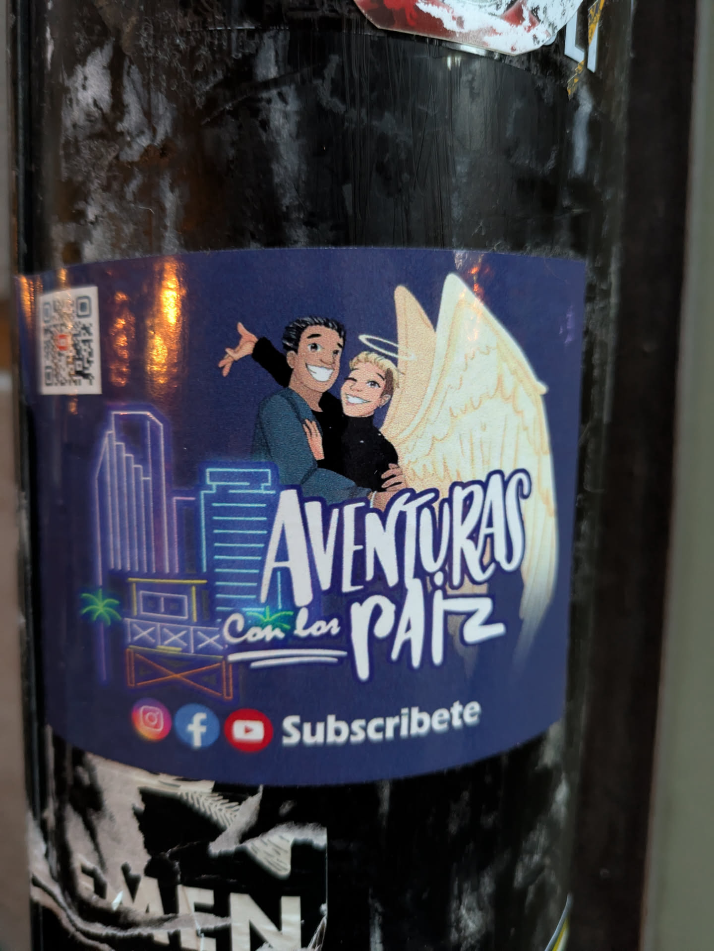 The sticker features a cartoon illustration on a dark blue background. In the upper portion, a smiling couple is depicted. The person on the left has dark hair and is wearing a teal shirt, with their arm around the person on the right. The person on the right has blonde hair and a halo above their head, suggesting an angelic theme. Large, light yellow angel wings are spread behind them. Below the couple, a stylized cityscape with neon blue outlines of buildings is shown, along with palm trees and a wooden structure. The text 'AVENTURAS Con los PAIZ' is prominently displayed in white, bubbly lettering. At the bottom, social media icons for Instagram, Facebook, and YouTube are visible, followed by the text 'Subscribete'. A QR code is located on the far left of the sticker.