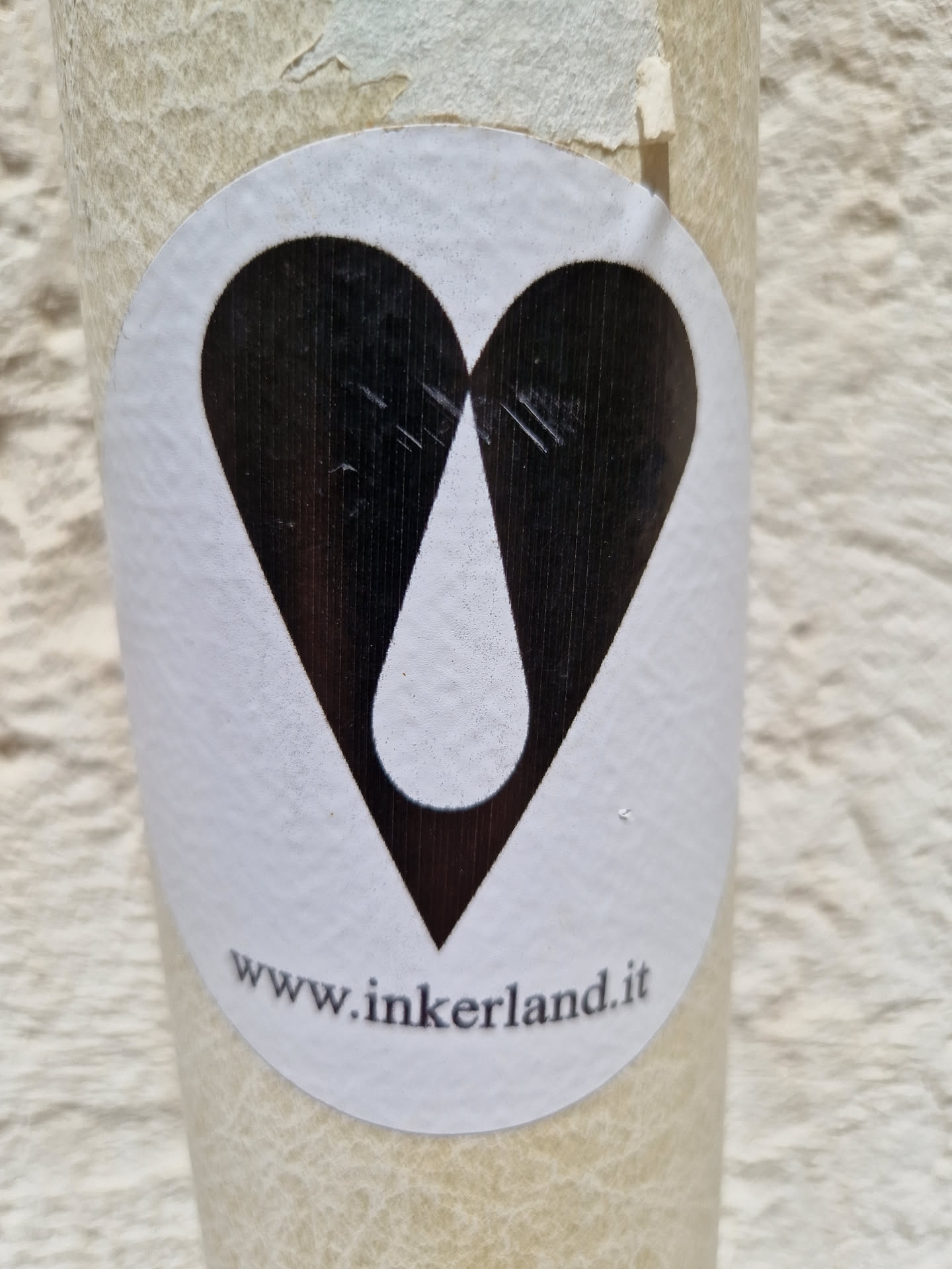 White round sticker on a beige surface with a black heart shape containing a white teardrop in its center. At the bottom, a website address is visible: www.inkerland.it