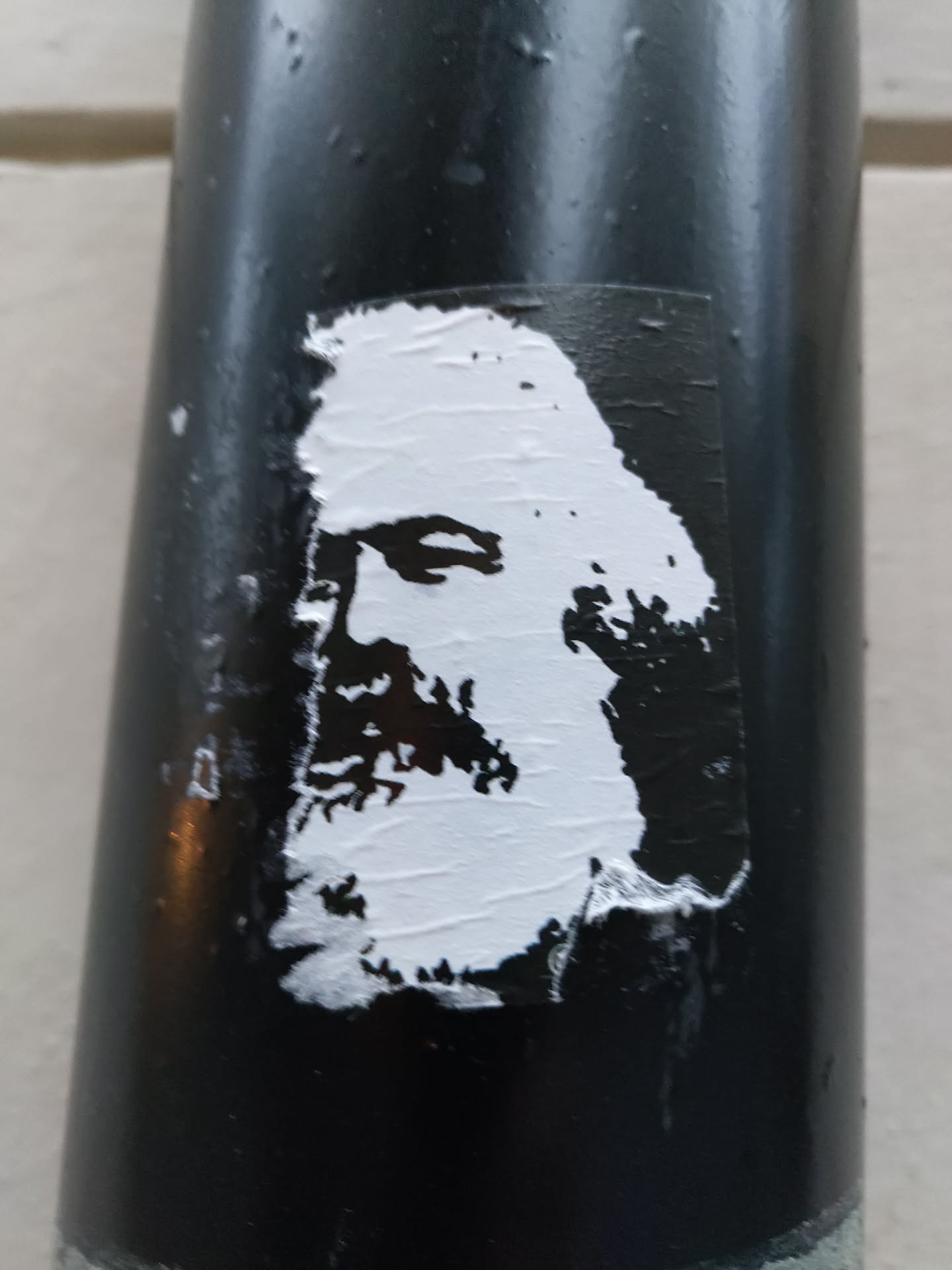 A black and white sticker featuring a stylized portrait of Karl Marx. The sticker is affixed to a dark-colored cylindrical surface, possibly a pole or pipe.