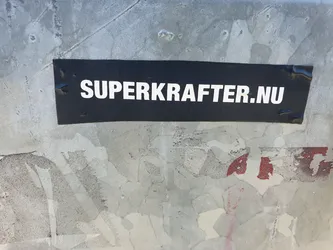 A black rectangular sticker with the text "SUPERKRAFTER.NU" in white uppercase letters is affixed to a weathered gray surface. The surface shows signs of wear and tear, with peeling paint and scratches.