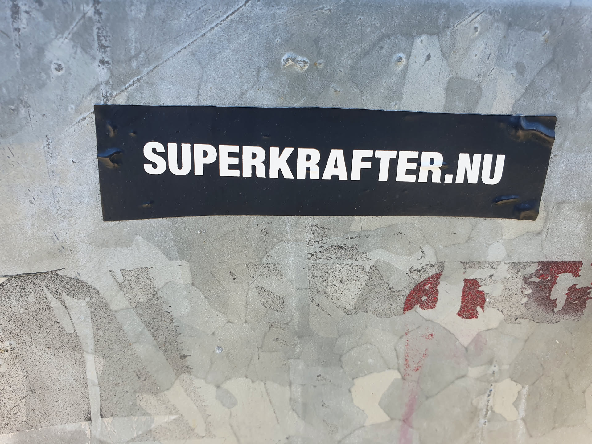 A black rectangular sticker with the text "SUPERKRAFTER.NU" in white uppercase letters is affixed to a weathered gray surface. The surface shows signs of wear and tear, with peeling paint and scratches.