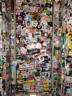 This image shows a wall completely covered in stickers of varying sizes, colors, and designs.  Many have text, images, and logos. The stickers create a complex and vibrant collage. It appears to be located outdoors, likely in a public space. Some stickers contain text, such as "ONE LOVE" and "Mook Life", but not all text is legible.