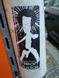 Black and white sticker with a stylized figure reminiscent of religious iconography. The figure stands within a sunburst, with the text "Neon Judas" at the bottom. The sticker is affixed to a weathered pole with cracked paint.