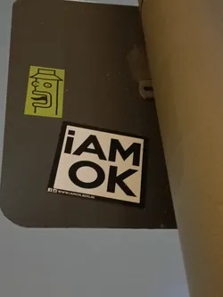 A black and white sticker with the text "i am ok" in bold capital letters.  There is a smaller, yellow sticker of a stylized face above it. A social media link is included at the bottom.