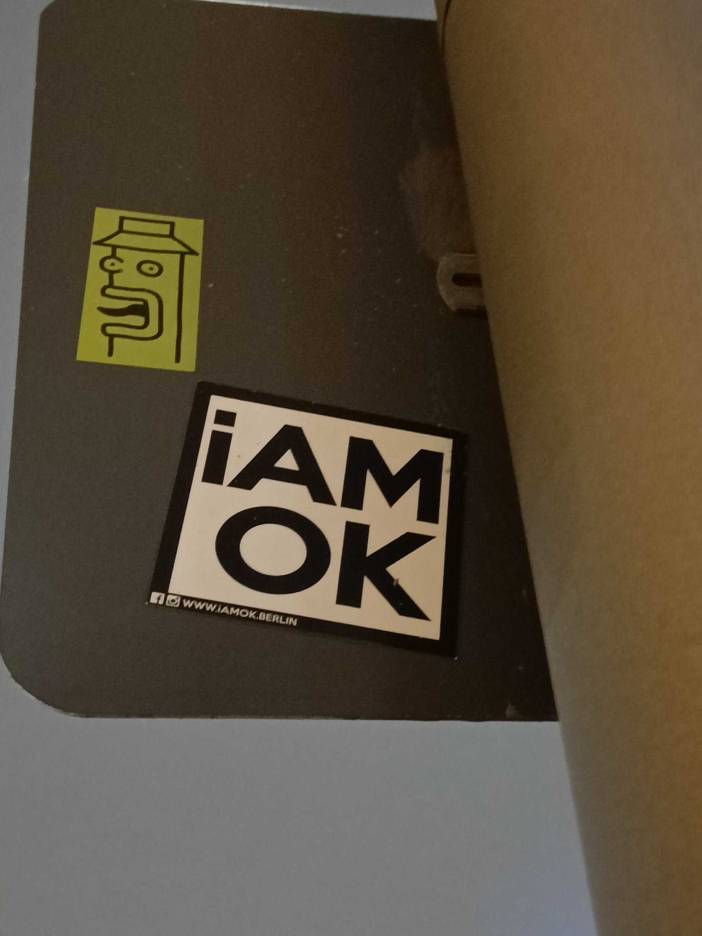 A black and white sticker with the text "i am ok" in bold capital letters.  There is a smaller, yellow sticker of a stylized face above it. A social media link is included at the bottom.