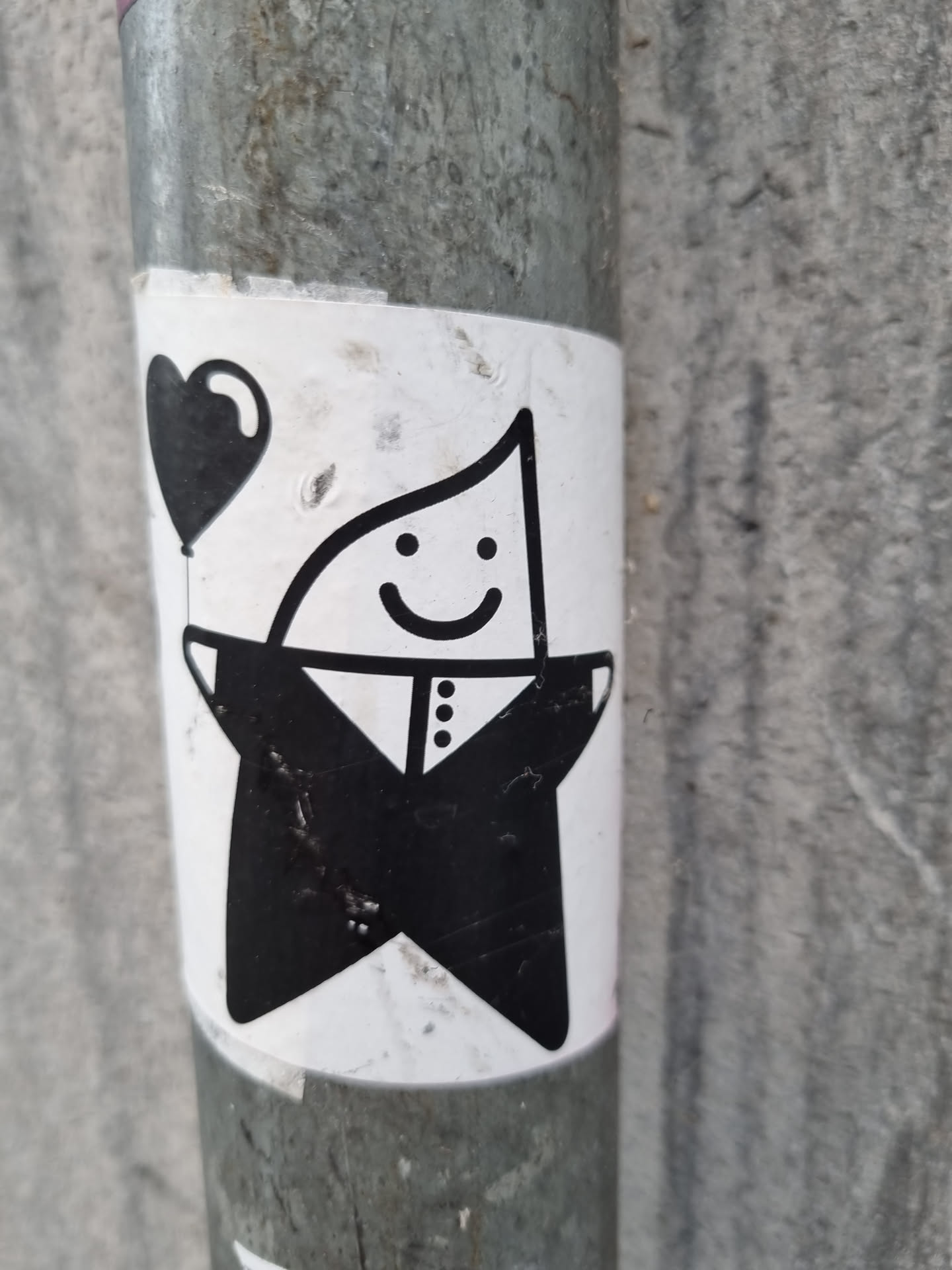 A black and white sticker featuring a cute, smiling, star-shaped character in a tuxedo holding a heart-shaped balloon. The sticker is affixed to a metal pole. The style is minimalist and charming.