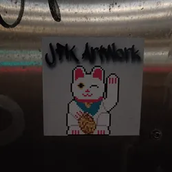 A pixel art sticker featuring a Maneki-neko (Japanese beckoning cat) holding a coin. The sticker has the text "JPK Artwork" printed on it. The cat is white with pink ears and paws, and is depicted in a pixelated style.