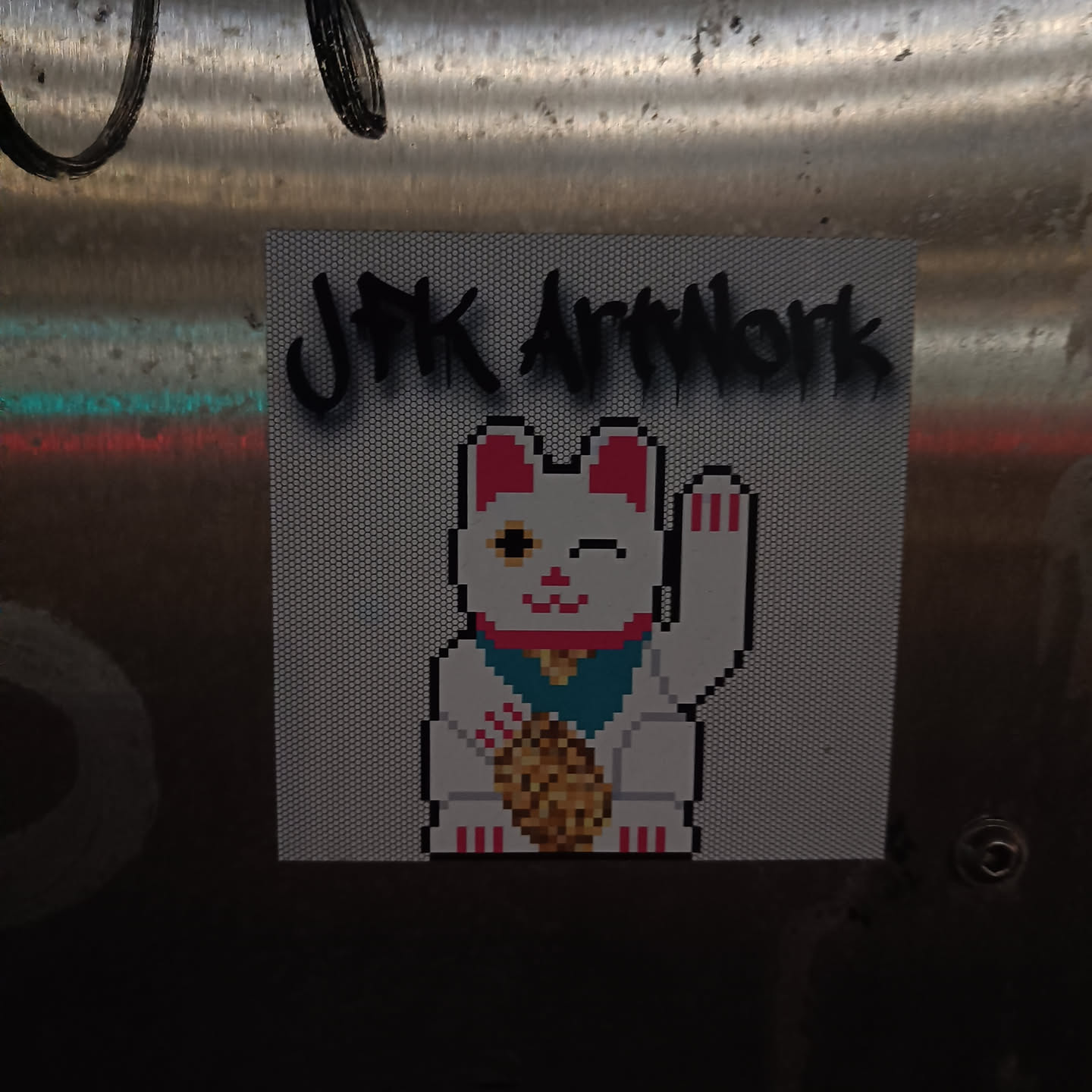 A pixel art sticker featuring a Maneki-neko (Japanese beckoning cat) holding a coin. The sticker has the text "JPK Artwork" printed on it. The cat is white with pink ears and paws, and is depicted in a pixelated style.