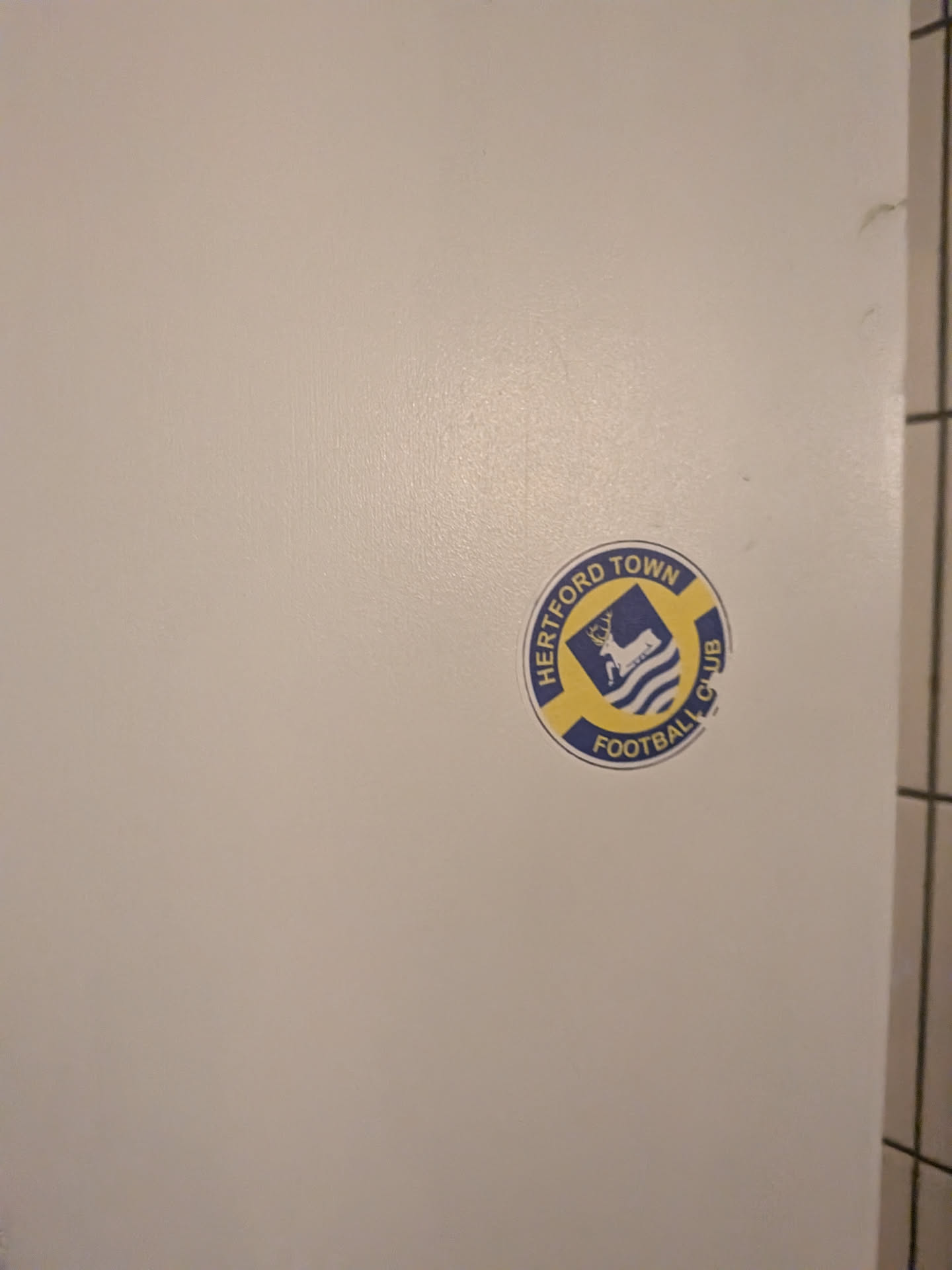 A circular sticker featuring the logo of Hertford Town Football Club. The design consists of a blue outer ring with yellow text that reads 'HERTFORD TOWN' at the top and 'FOOTBALL CLUB' at the bottom. The inner part of the logo is a blue shield shape, which contains a white stag (deer) standing on three white wavy lines, likely representing water. The overall color scheme is blue, yellow, and white.