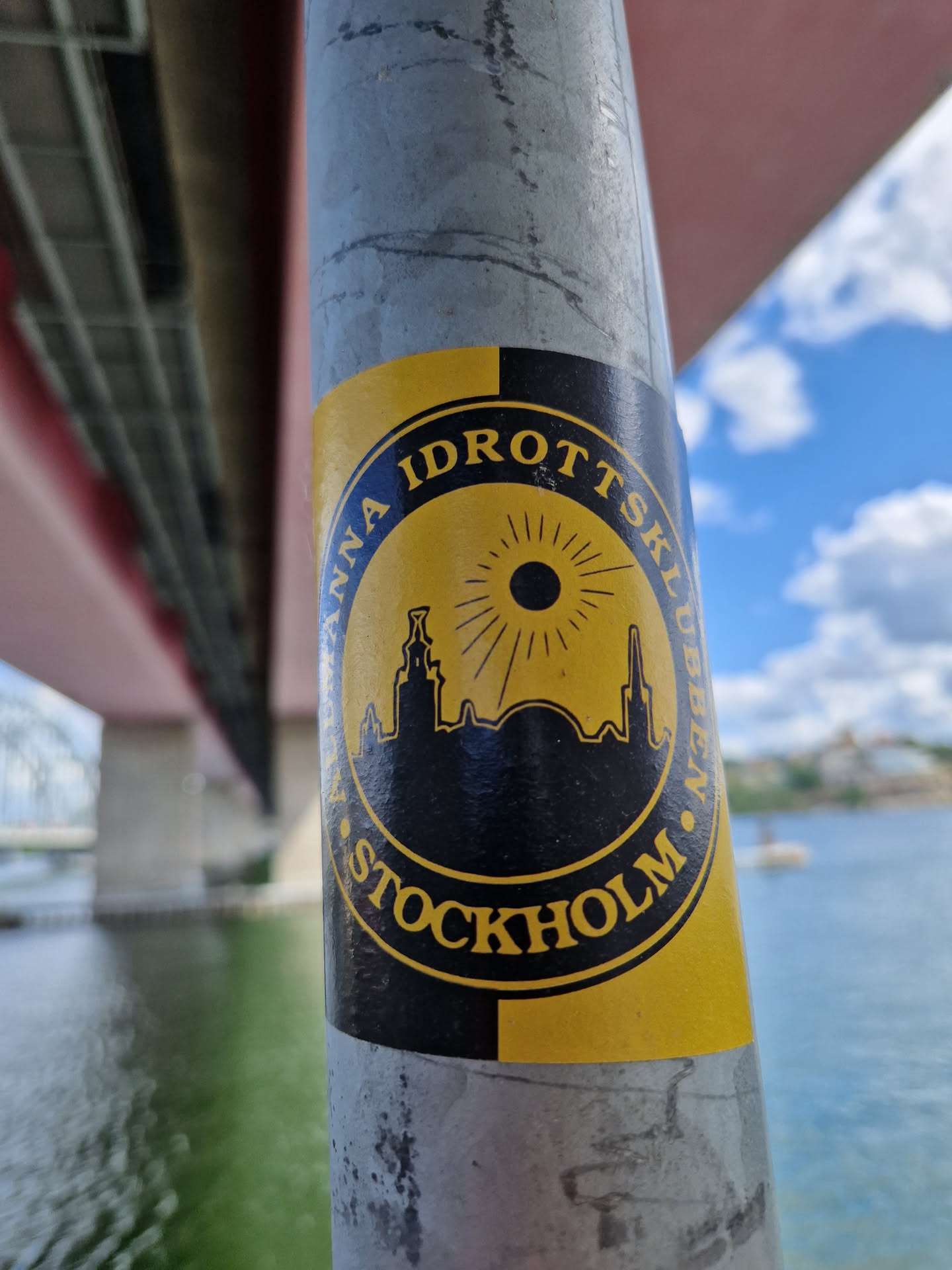 A yellow and black circular sticker featuring a stylized cityscape of Stockholm. The text around the circle reads "Anna Idrottsklubben Stockholm". The design is simple yet striking, with a sunburst graphic behind the city skyline. The sticker is affixed to a metal pole.