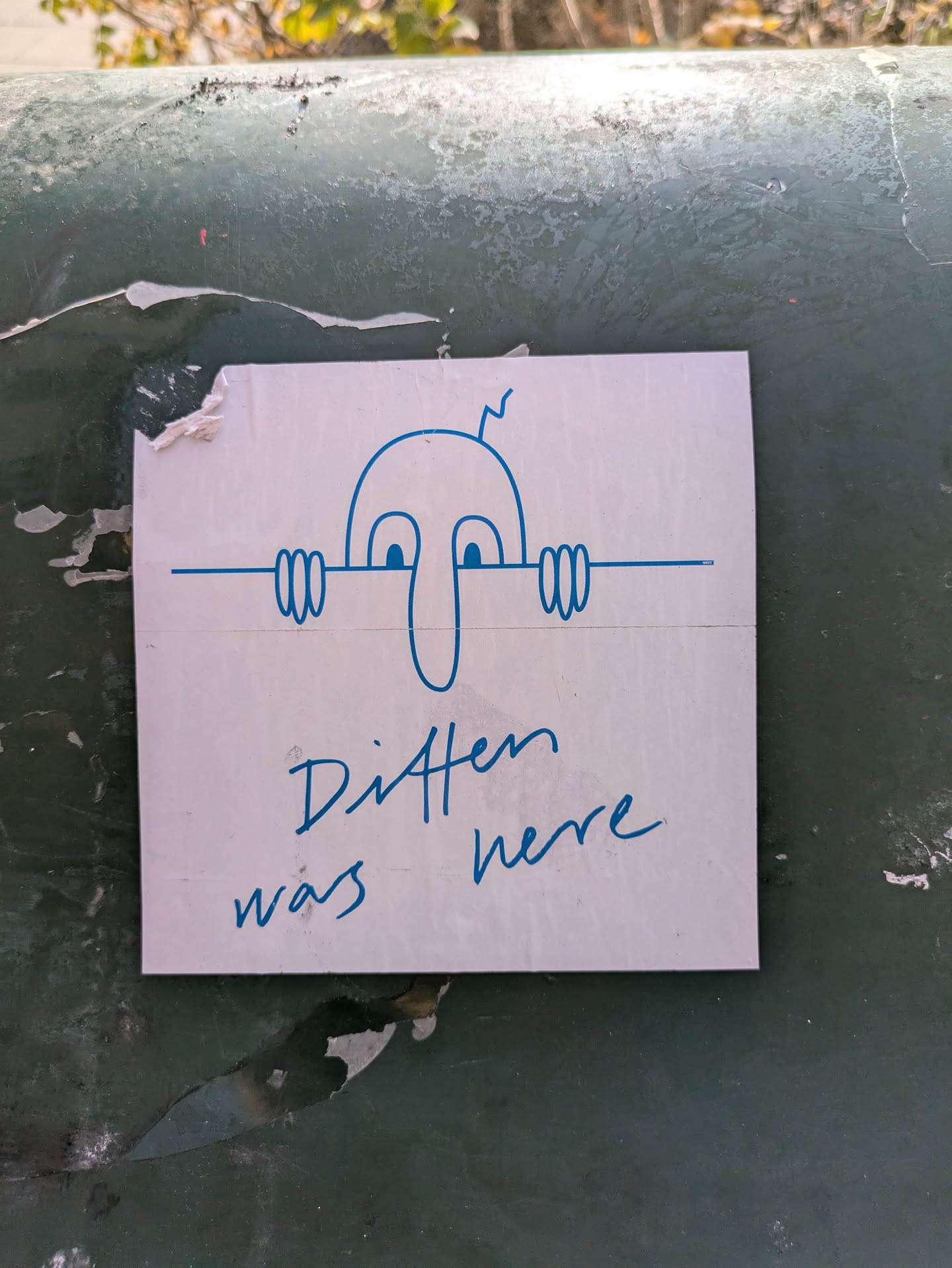 A white sticker with a simple drawing of a face peeking over a line. Blue ink is used for the drawing and the text. The text on the sticker says "Ditten was here".
