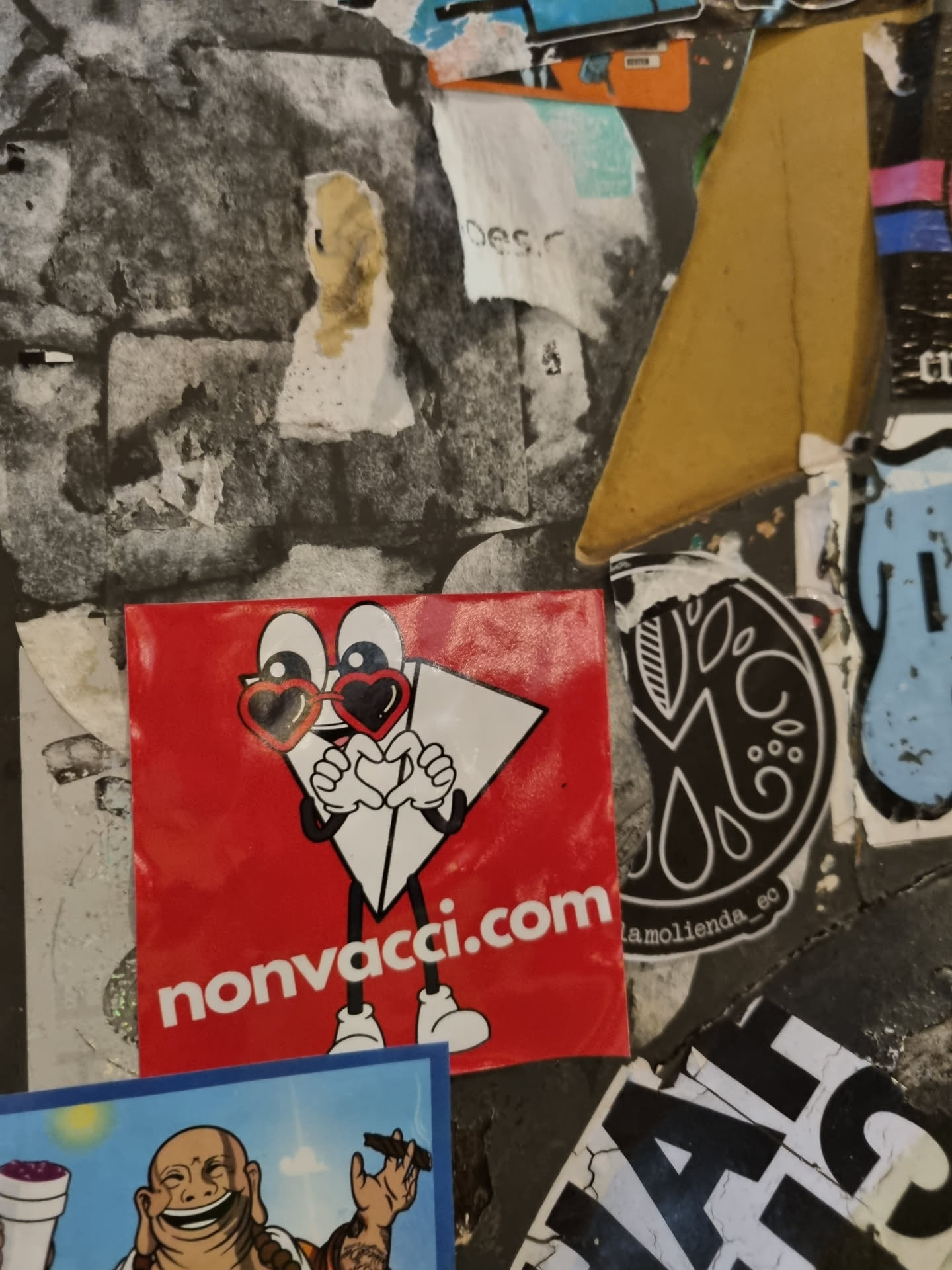 A red square sticker features a cartoon character resembling a paper airplane with heart-shaped sunglasses. The character is holding its hands in a heart shape. The website nonvacci.com is printed at the bottom. The sticker is affixed to a wall amongst other stickers.