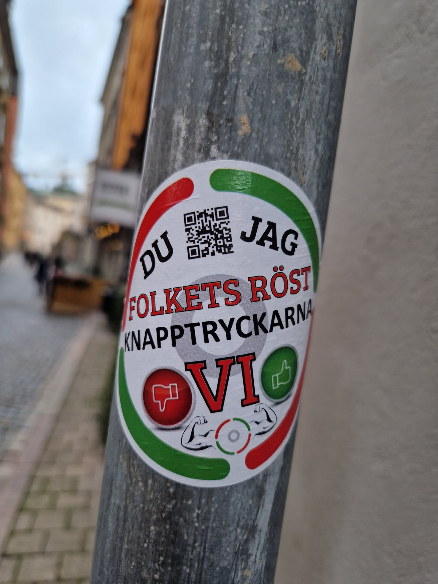 A sticker with a QR code, text in Swedish, and a design featuring thumbs up and down, along with images of biceps. The text translates to "The People's Voice - The Button Pushers VI".