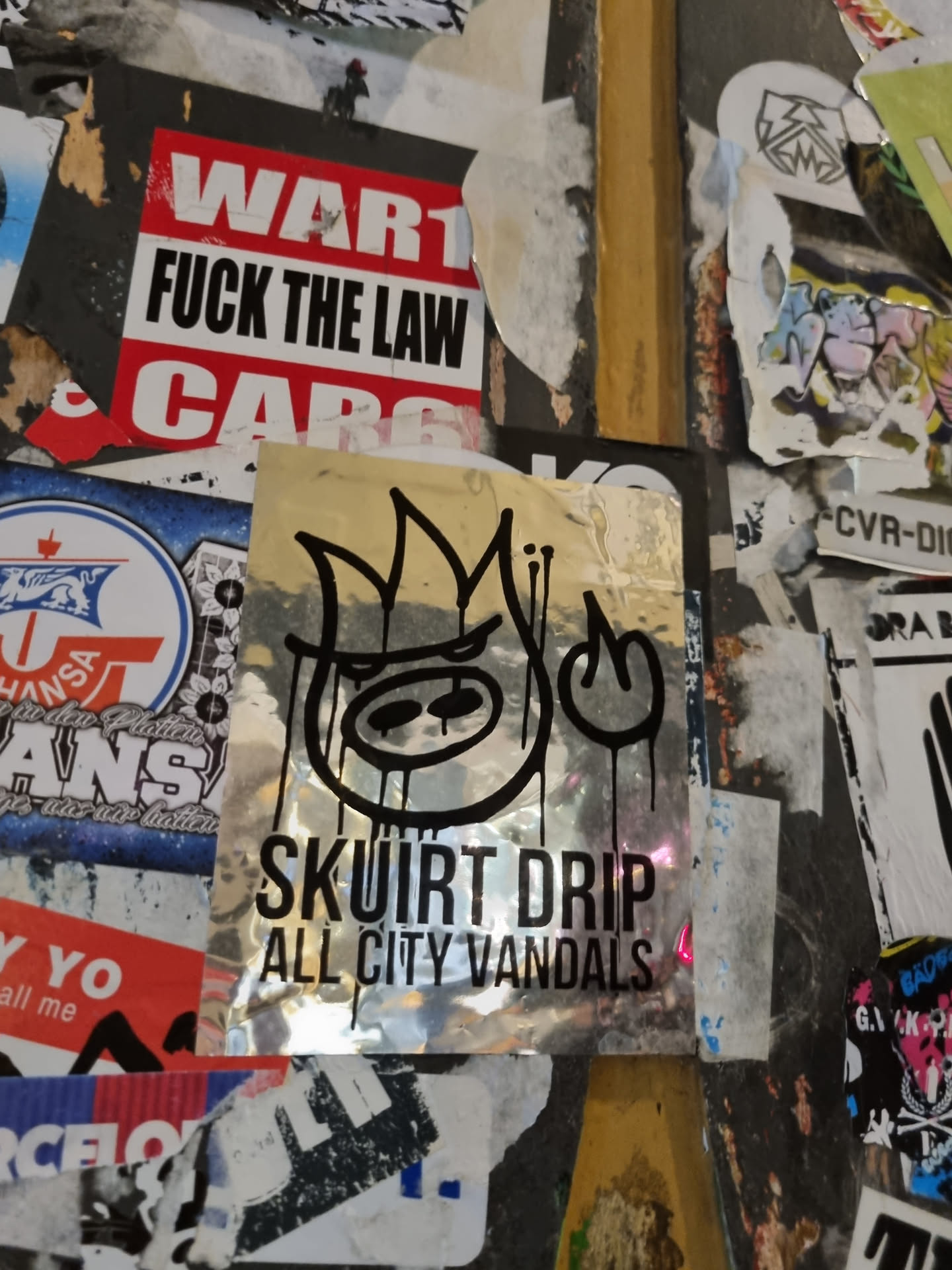 A silver sticker with a black graphic and text. The text says "Skuirt Drip" over "All City Vandals". The graphic appears to be a stylized face with flames.