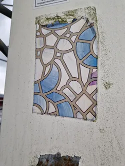 A weathered sticker featuring an abstract design reminiscent of stained glass. The sticker is predominantly beige and blue, with a geometric pattern of interconnected shapes. The sticker is affixed to a light beige surface, showing signs of wear and tear. 