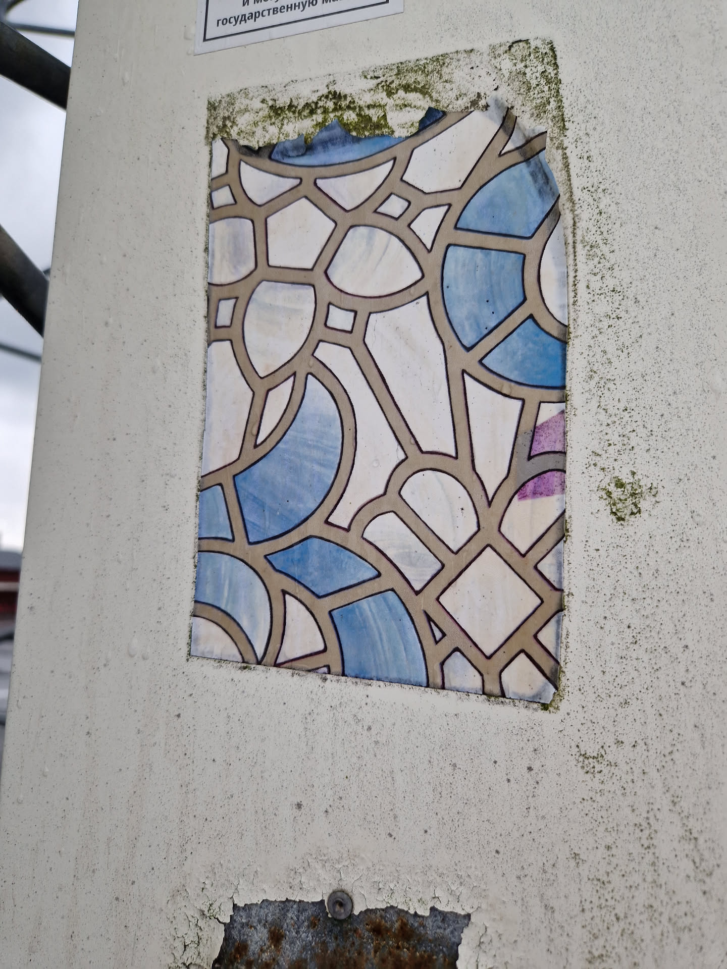 A weathered sticker featuring an abstract design reminiscent of stained glass. The sticker is predominantly beige and blue, with a geometric pattern of interconnected shapes. The sticker is affixed to a light beige surface, showing signs of wear and tear. 