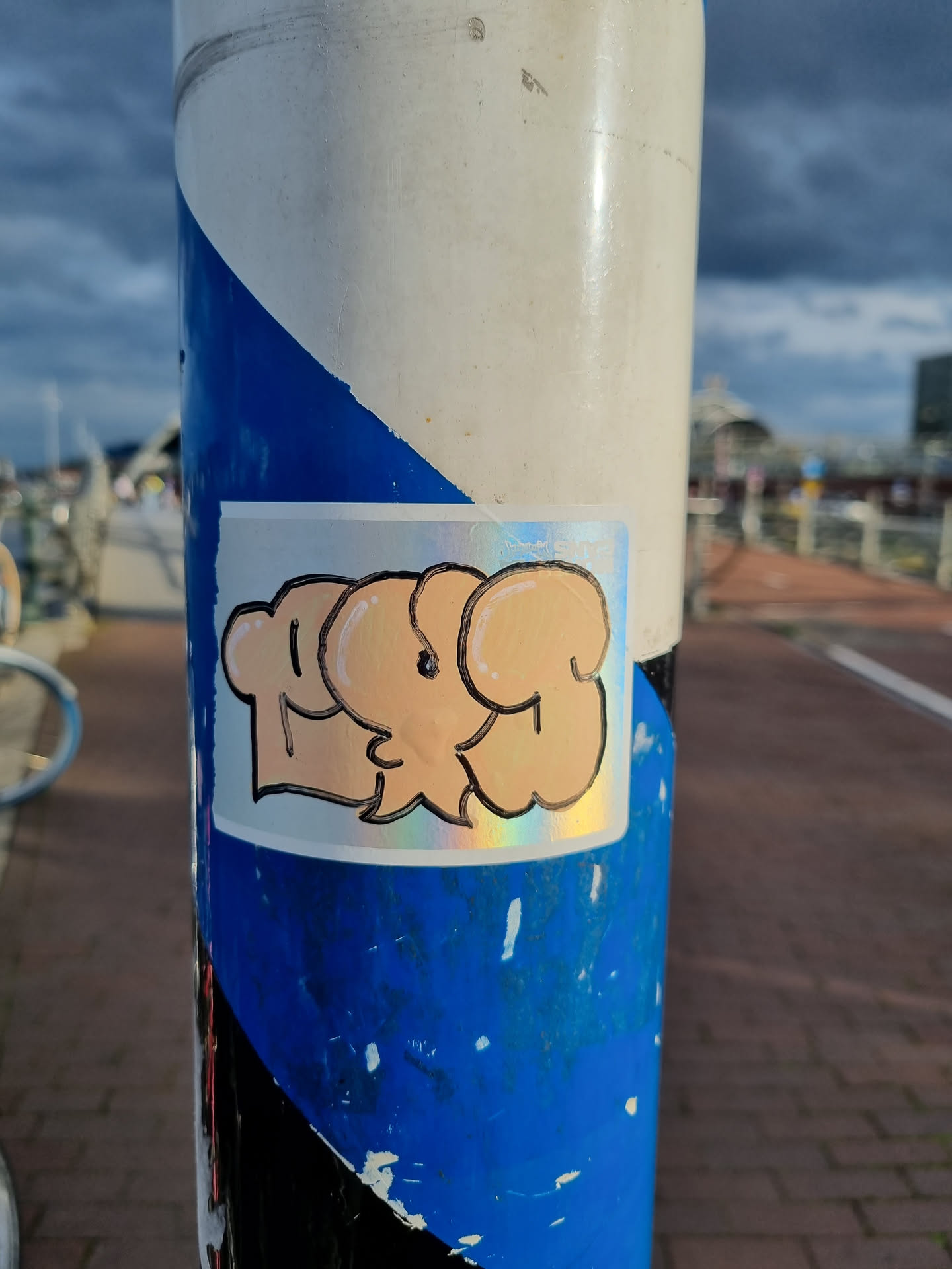 A holographic sticker featuring a unique design of three abstract figures in a light peach color. The sticker is affixed to a weathered pole with blue and white stripes. 