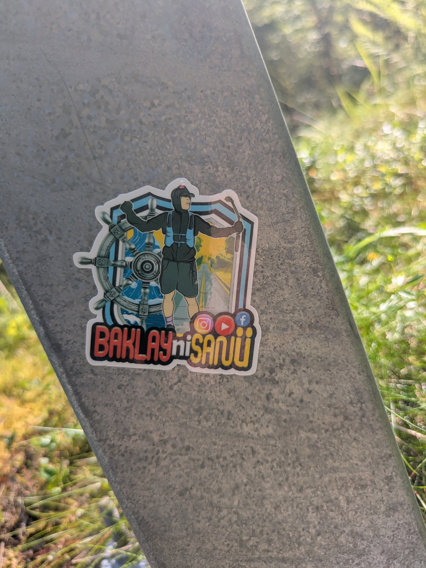 The sticker depicts a stylized character, possibly an adventurer or runner, wearing a cap, dark jacket with a blue hydration vest, and shorts. The character stands in front of a dynamic background split into two main elements: on the left, a large grey ship's steering wheel with blue waves behind it, suggesting sea or nautical themes; on the right, a winding road lined with trees under a bright sky, indicating travel or journey. Below the character, there are three distinct social media logos: Instagram (orange square), YouTube (red play button), and Facebook (blue 'f' icon). The text 'BAKLAY ni SANUÜ' is prominently displayed in bold, two-tone lettering (red/orange and yellow). The entire design has a white outline, making it stand out against the metallic surface it's affixed to.