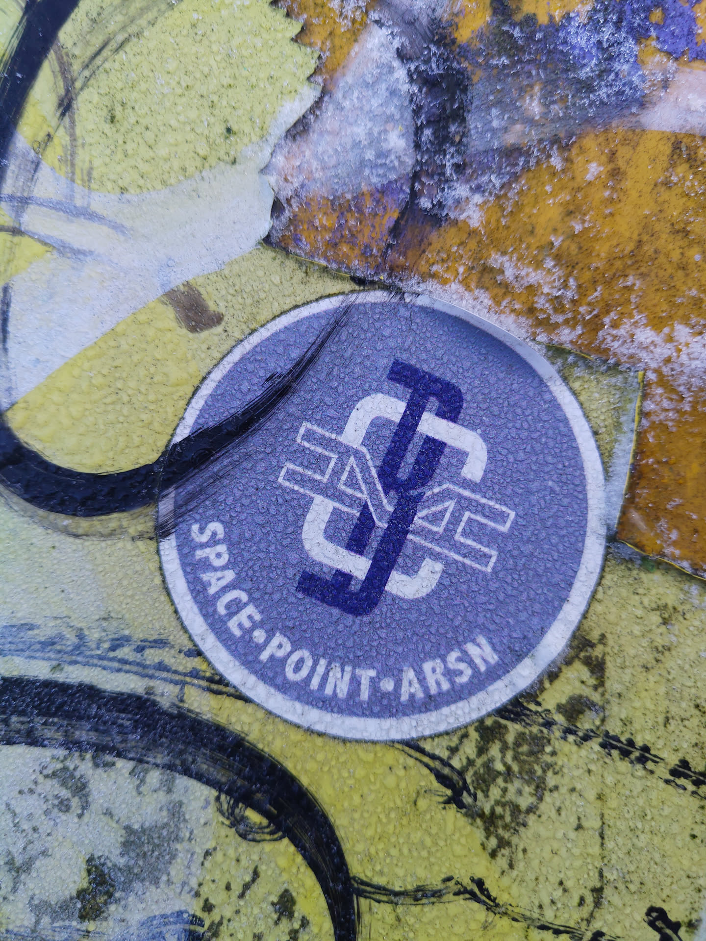 A round sticker with a logo featuring overlapping letters 'S', 'P', and 'A', possibly an abbreviation for Space Point ArsN. The sticker is light purple with white text. It appears to be affixed to a wall with other graffiti.