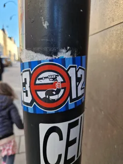 A sticker with a protest theme against police brutality. It features a symbol of a person raising a fist in protest inside a circle with a slash through it, combined with the numbers 13012. The sticker is affixed to a pole.