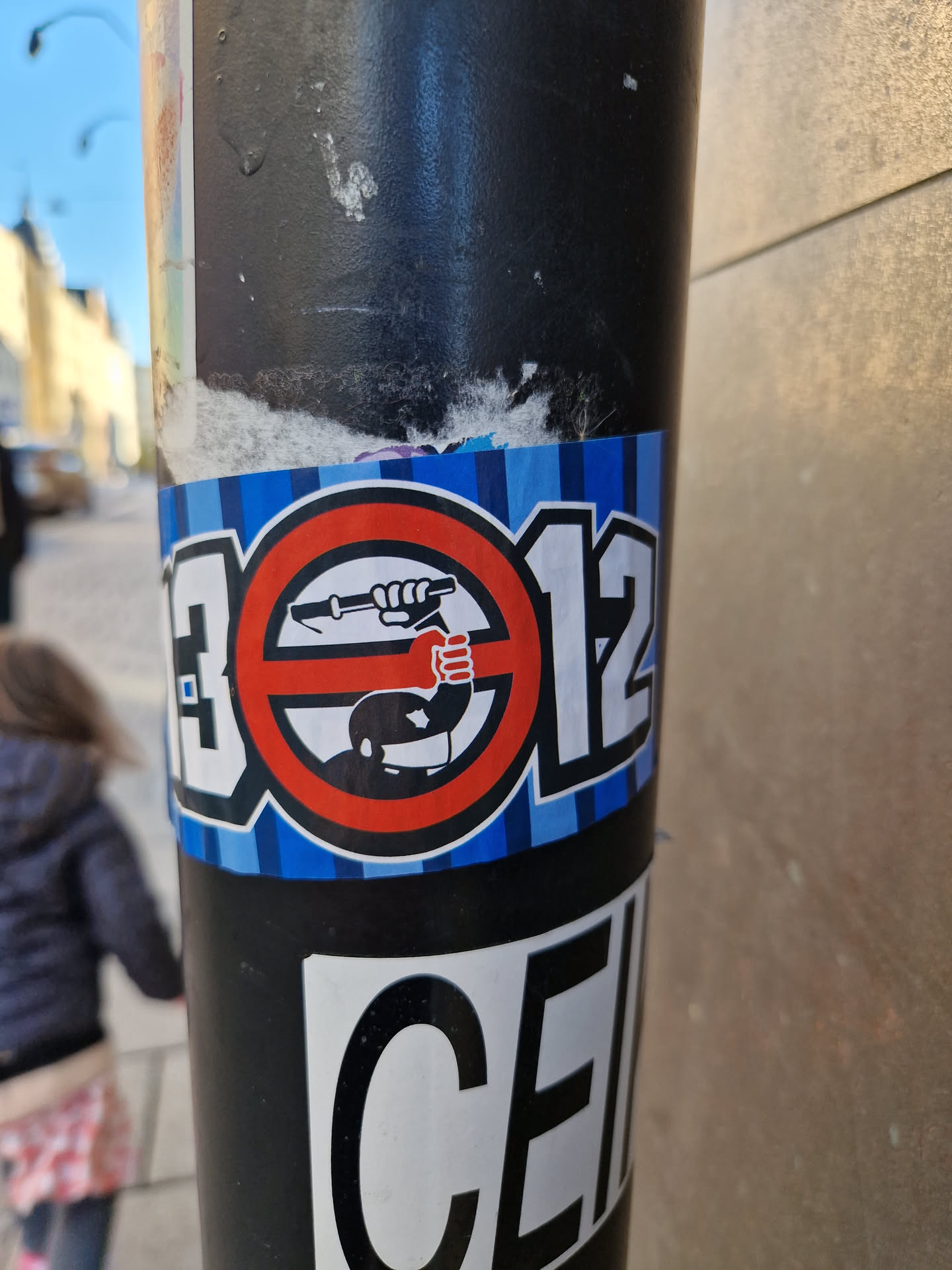 A sticker with a protest theme against police brutality. It features a symbol of a person raising a fist in protest inside a circle with a slash through it, combined with the numbers 13012. The sticker is affixed to a pole.