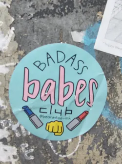 Round sticker with the text "Badass babes club" in pink and black on a light blue background. It also features illustrations of lipsticks and a fist.