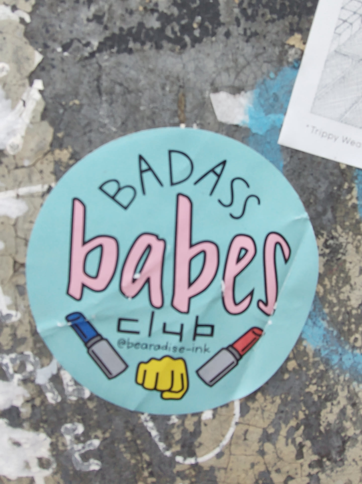 Round sticker with the text "Badass babes club" in pink and black on a light blue background. It also features illustrations of lipsticks and a fist.