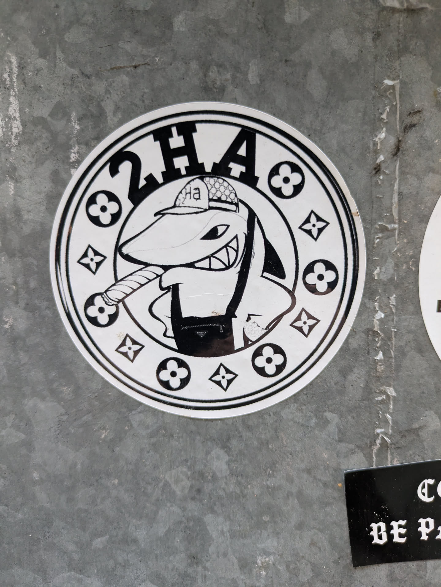 Black and white sticker of a shark wearing a hat and smoking a cigar. The design is surrounded by a circular border with repeating Louis Vuitton-style flower patterns and the text "2HA".