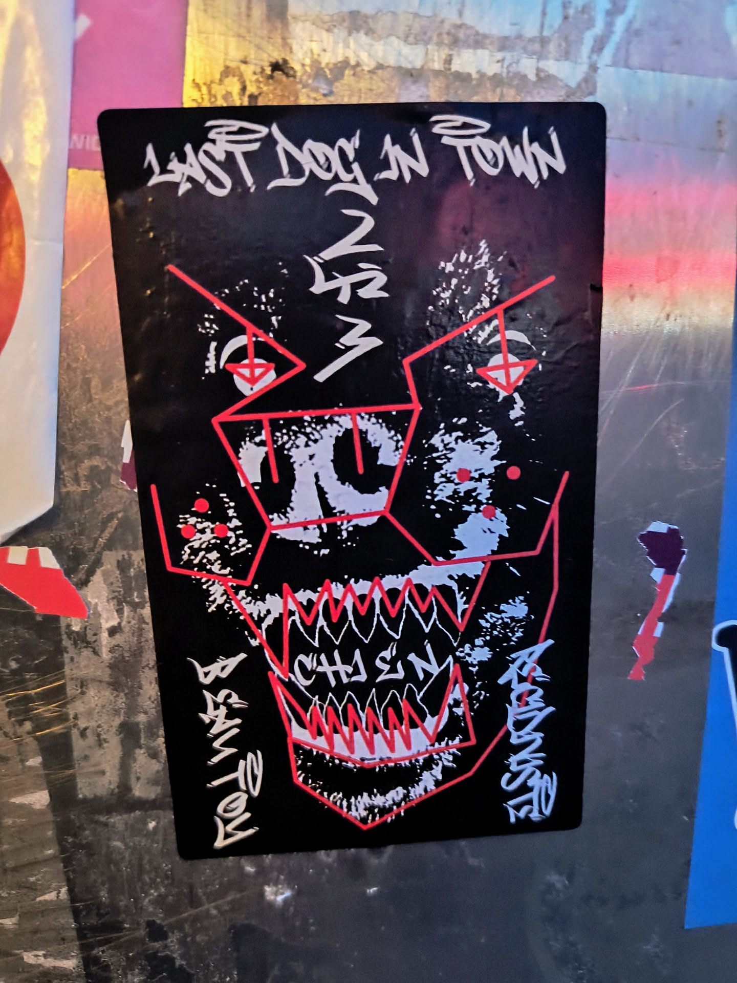 A black sticker shows an abstract depiction of a dog's face with red geometric lines and jagged white teeth. The text "LAST DOG IN TOWN" is at the top, with illegible artist signature on the bottom.