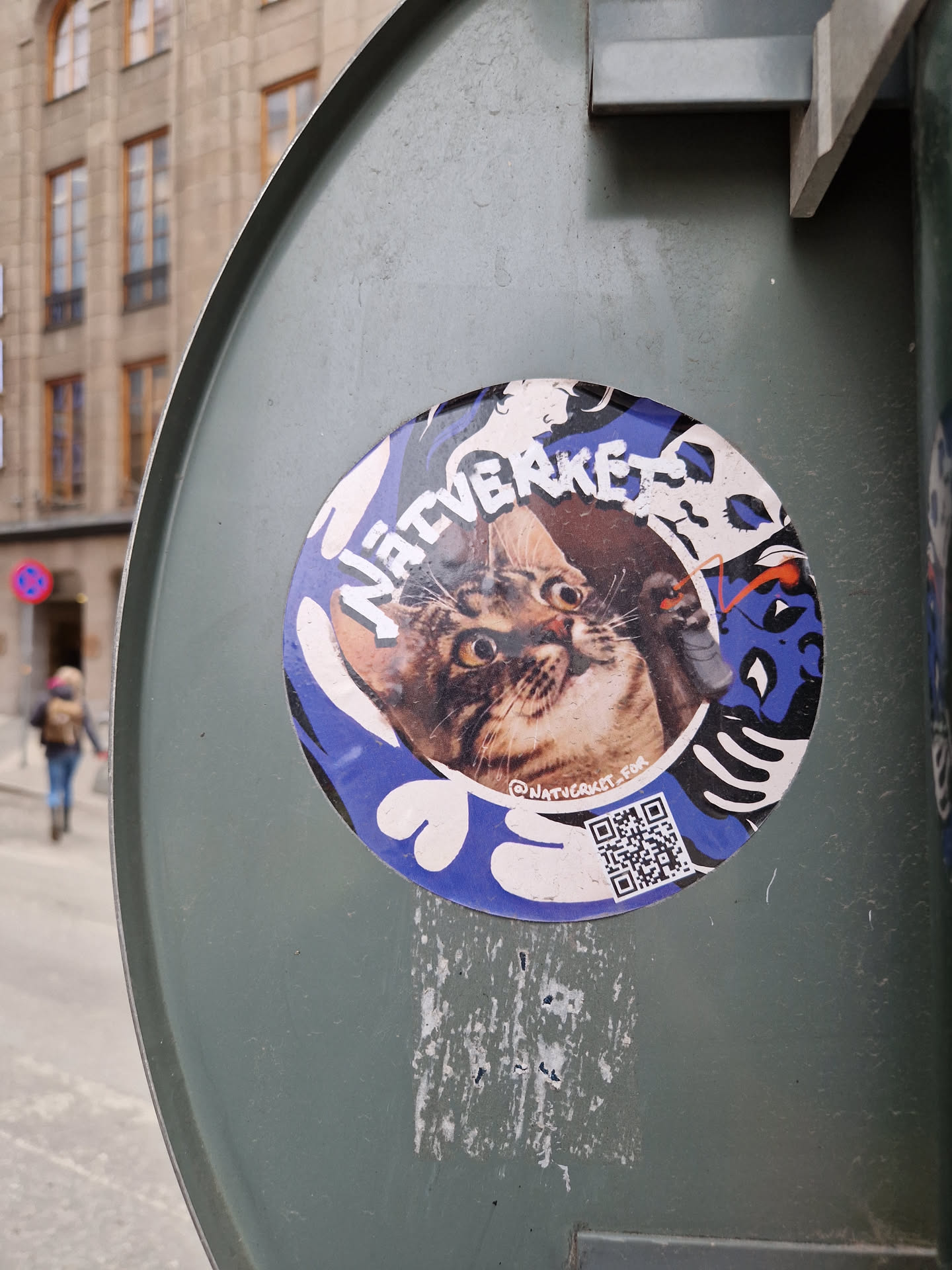 A round sticker featuring a cat's face and the words "Native Rocket." The design is vibrant, with blue, white, and other colors. A QR code is also present at the bottom. The sticker is affixed to a weathered gray metal surface.