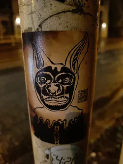 A sticker featuring a stylized, almost demonic-looking dog or similar creature with large ears. The art style is dark and graphic, with black ink on what appears to be a beige or tan background. The sticker seems to be affixed to a weathered pole, and a small QR code is visible on the right side of the sticker. Another sticker is partially visible below this one.