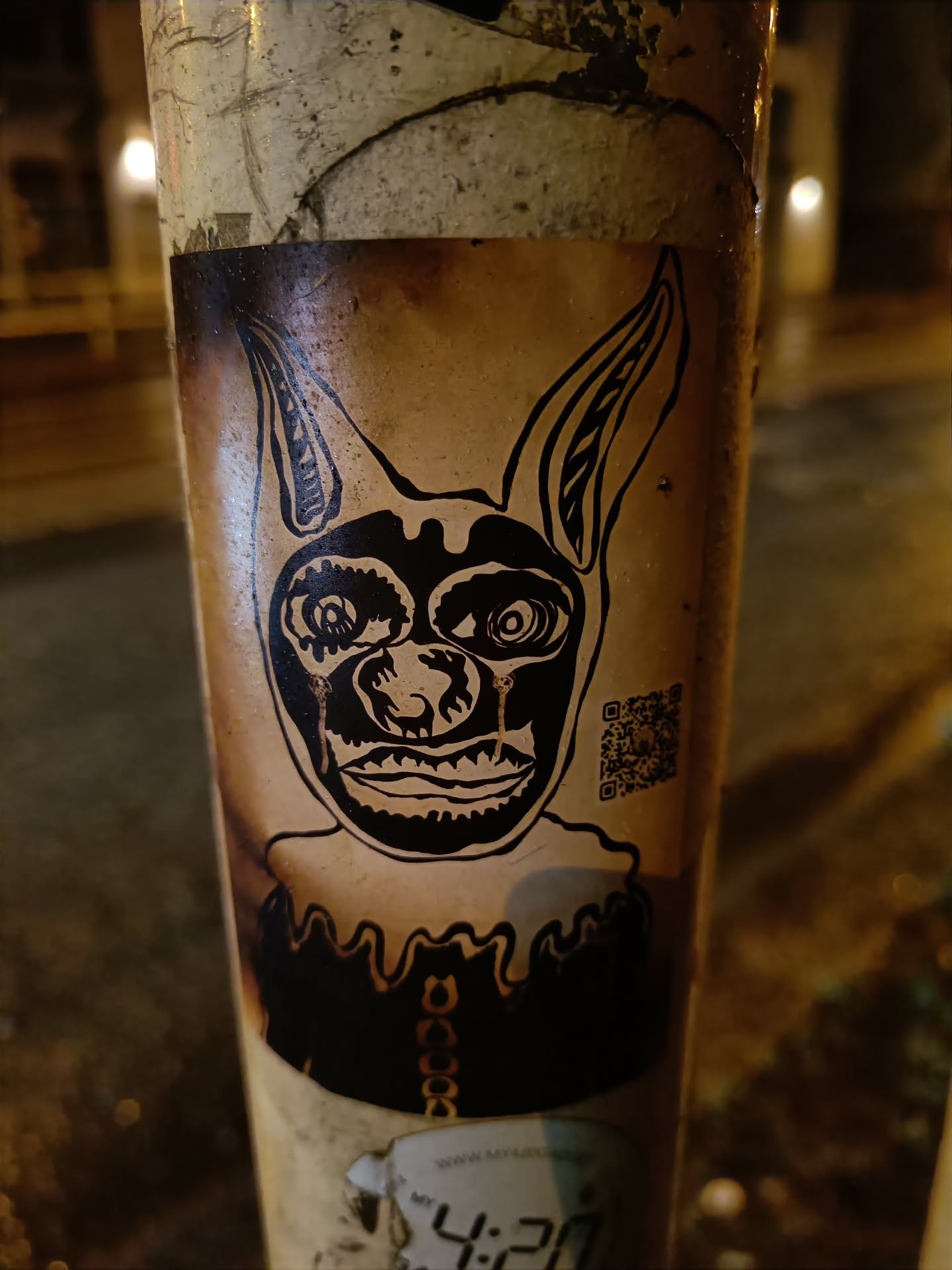 A sticker featuring a stylized, almost demonic-looking dog or similar creature with large ears. The art style is dark and graphic, with black ink on what appears to be a beige or tan background. The sticker seems to be affixed to a weathered pole, and a small QR code is visible on the right side of the sticker. Another sticker is partially visible below this one.
