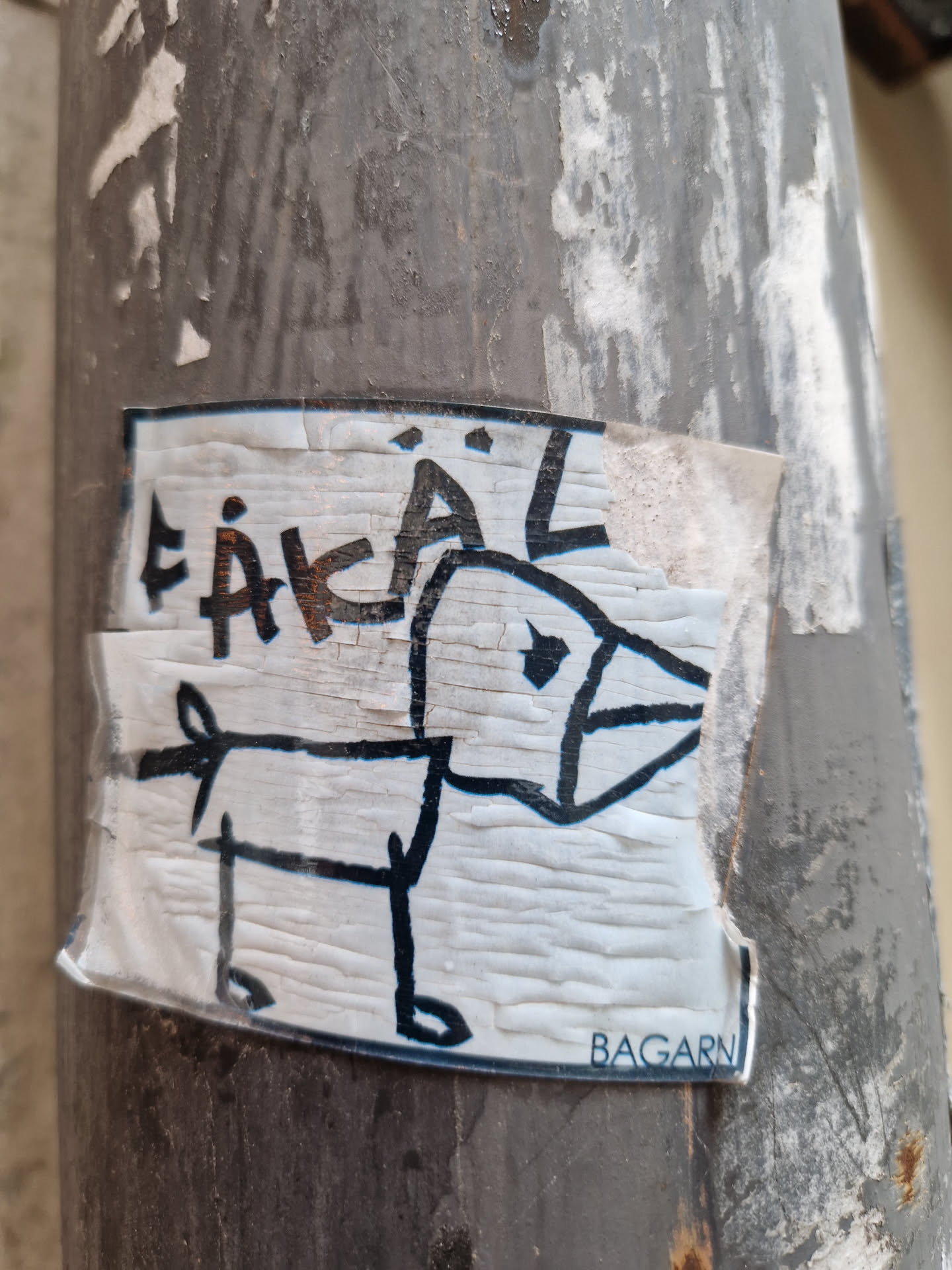 This sticker features a simple drawing of a dog, reminiscent of a child's artwork. The word "ÄKAL" is written above the dog in a stylized manner.  The sticker is affixed to a weathered surface, and shows signs of wear and tear. The word "BAGARN" is visible near the bottom.
