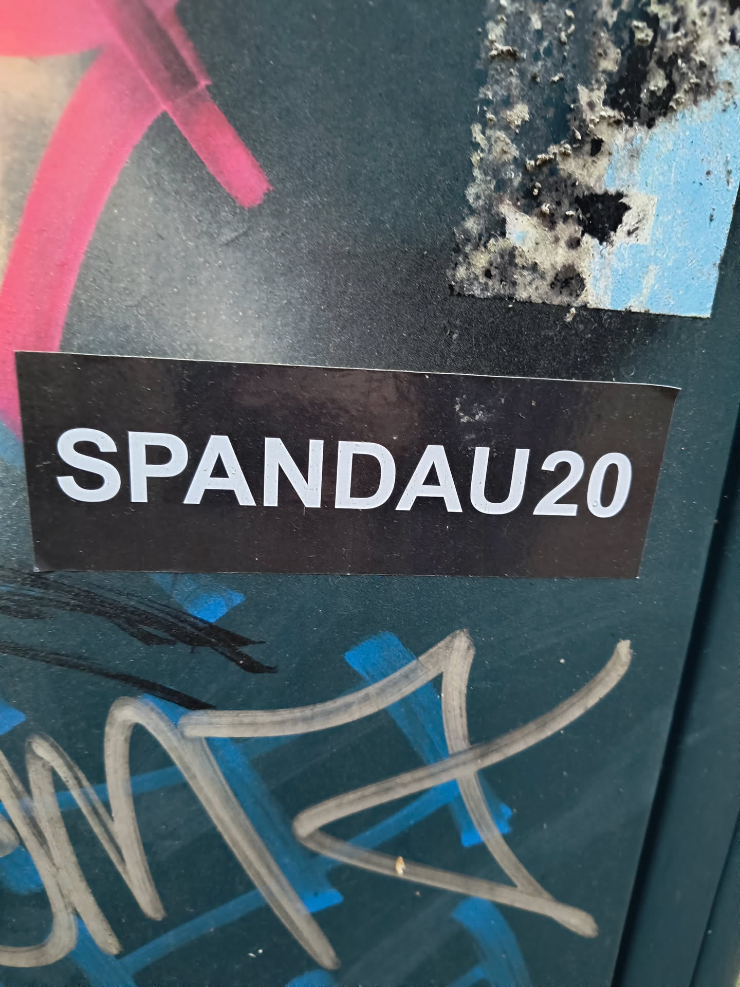 The sticker is rectangular with a black background and white, sans-serif text that reads 'SPANDAU20'. It is positioned centrally in the image, slightly overlapping with some graffiti elements. The sticker appears to be made of a glossy material, and its edges are sharp. The background of the image shows signs of wear and tear, with other graffiti and peeling paint visible.