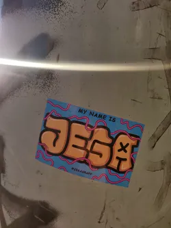 A sticker with a stylized name JESA in orange letters on a light blue background with pink lines. The text "My name is" is written on the top. The hashtag #JEGAGRAFF is visible at the bottom.