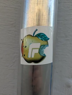 The sticker depicts a stylized apple, primarily yellow-green with a subtle reddish blush on its left side. A distinct bite mark is visible on the right side of the apple. A large, bold, white letter 'F' is integrated into the center of the apple design. A green leaf and a dark stem protrude from the top. On the left side, there are a few small, dark, circular elements that could represent seeds or small piercings. The sticker is affixed to a shiny, scratched metallic cylindrical surface.