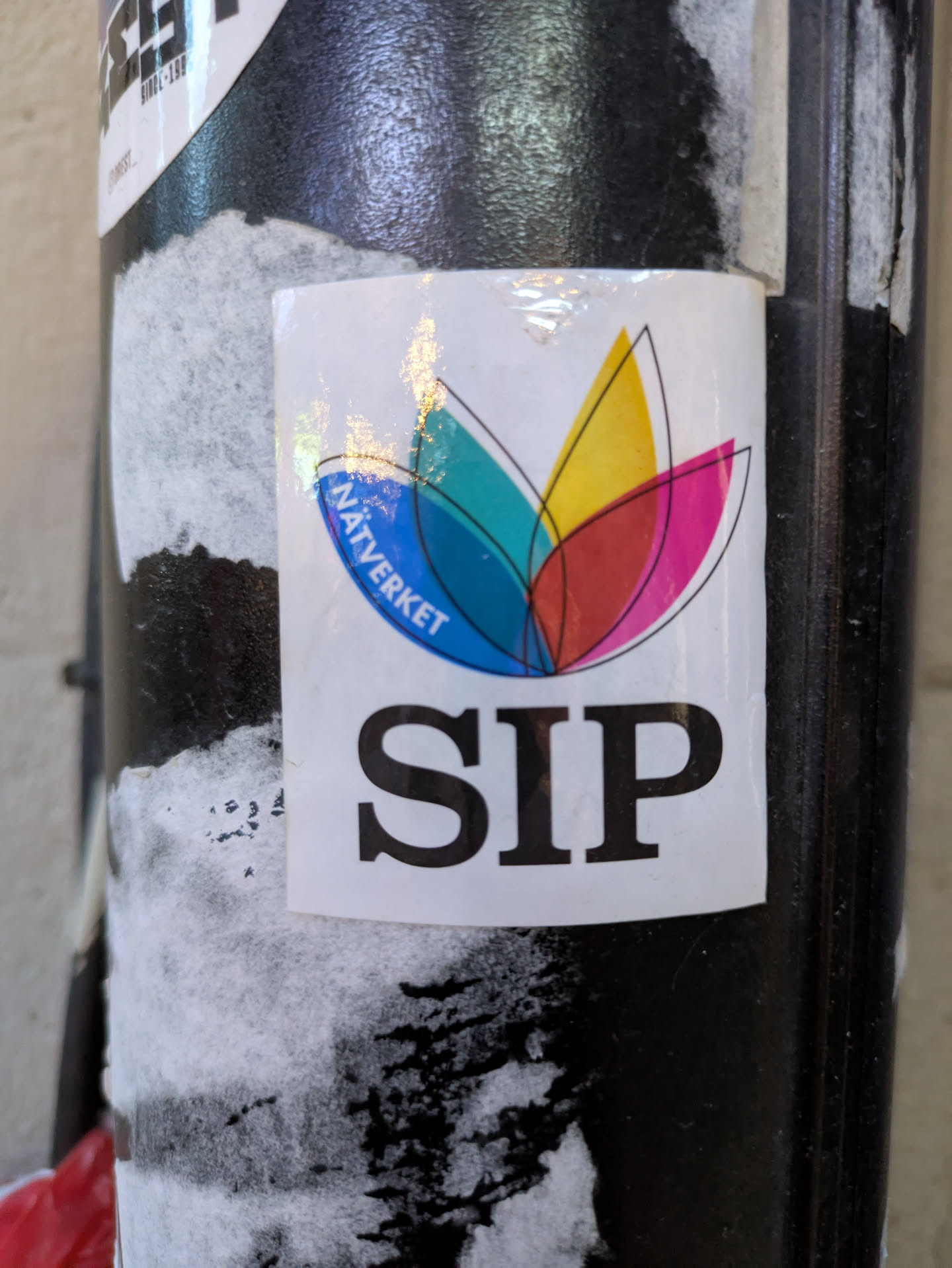 The sticker is primarily white with a central abstract design composed of four petal-like shapes in teal, yellow, red, and pink, outlined in black. Above the petals, within a curved blue segment, the word 'NÄTVERKET' is written in white text. Below the abstract design, the word 'SIP' is prominently displayed in large, black, sans-serif capital letters. The edges of the sticker appear slightly iridescent.