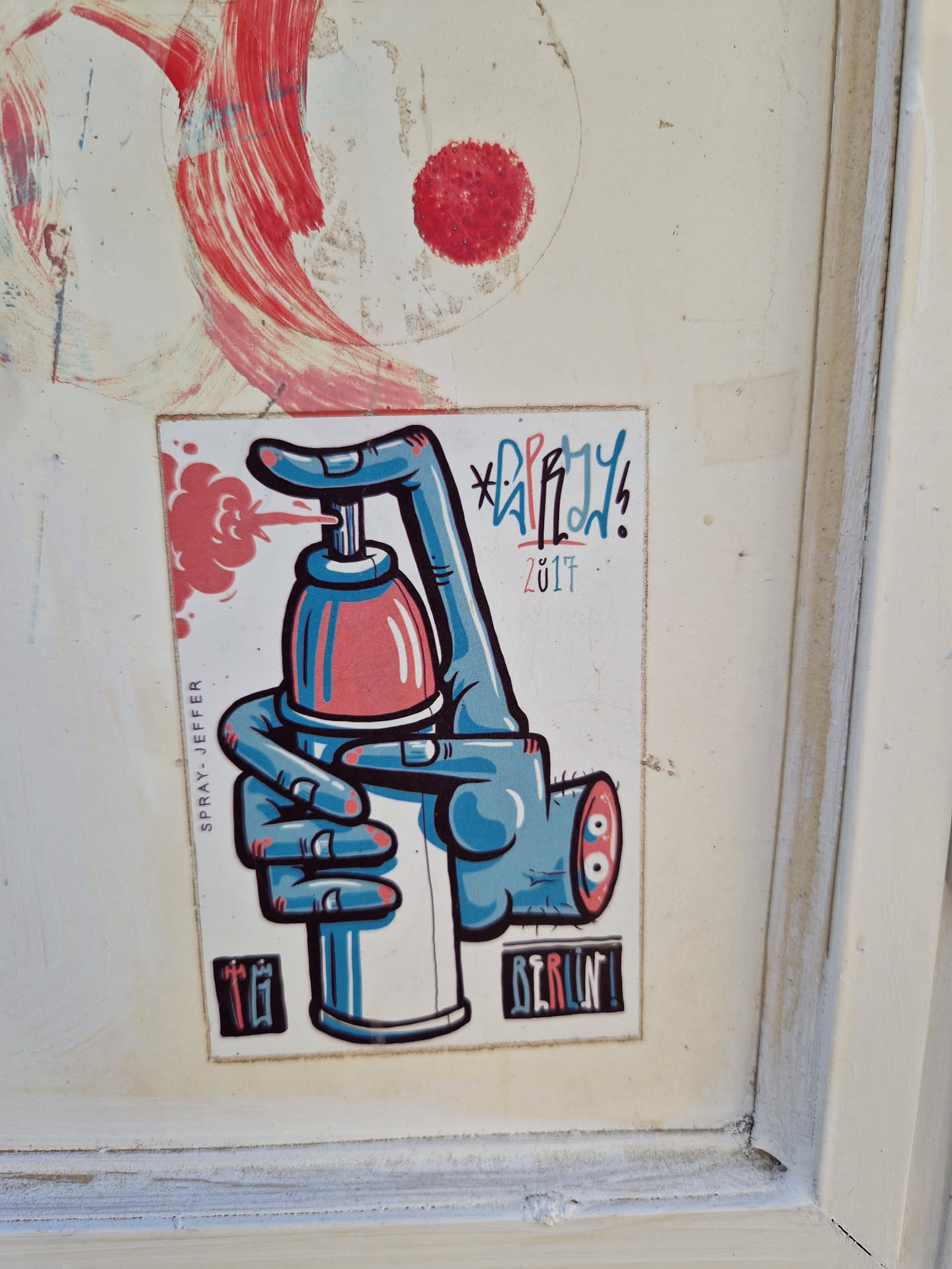 A sticker featuring a cartoon hand holding a spray can. The sticker is predominantly blue, red, and white. The artist's signature is visible at the bottom right corner, along with the word 'BERLIN!' indicating the location or theme. The year 2017 is printed in small font next to the artist's tag. The style is reminiscent of graffiti art, cartoonish and playful.