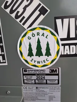 A round sticker with a white background bordered by alternating yellow and dark green triangular teeth forming an inner ring. In the center are three dark green silhouette-style pine trees, with the central tree being the tallest. Curving over the trees at the top is the word 'GÓRAL' in dark green sans-serif capital letters, and curving underneath is the word 'ŻYWIEC' in the same font and color.