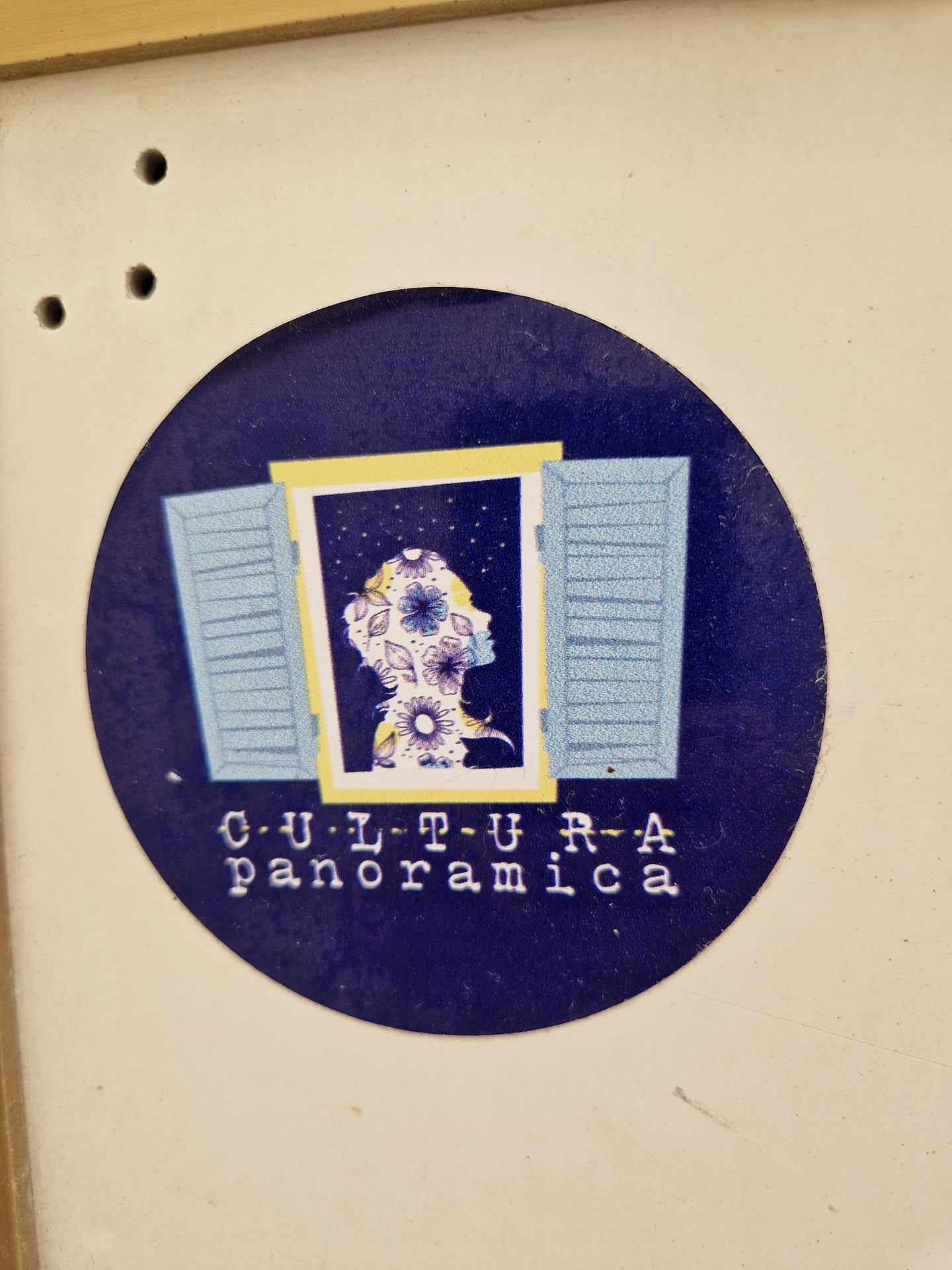 Round sticker featuring a stylized image of a woman's face behind a window with blue shutters. The background is dark blue and the text 'CULTURA panoramica' is displayed below the image.