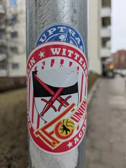 The sticker features the 1. FC Union Berlin academy logo. The logo is round with red and white stripes, and the text "BERLIN WITTENAU" is printed in red, surrounding the coat of arms of Wittenau, Berlin. Two swords are positioned behind the coat of arms. Below, the team's logo is situated within the 'FC' abbreviation. Lastly, the word "ACADEMY" is also written in red.