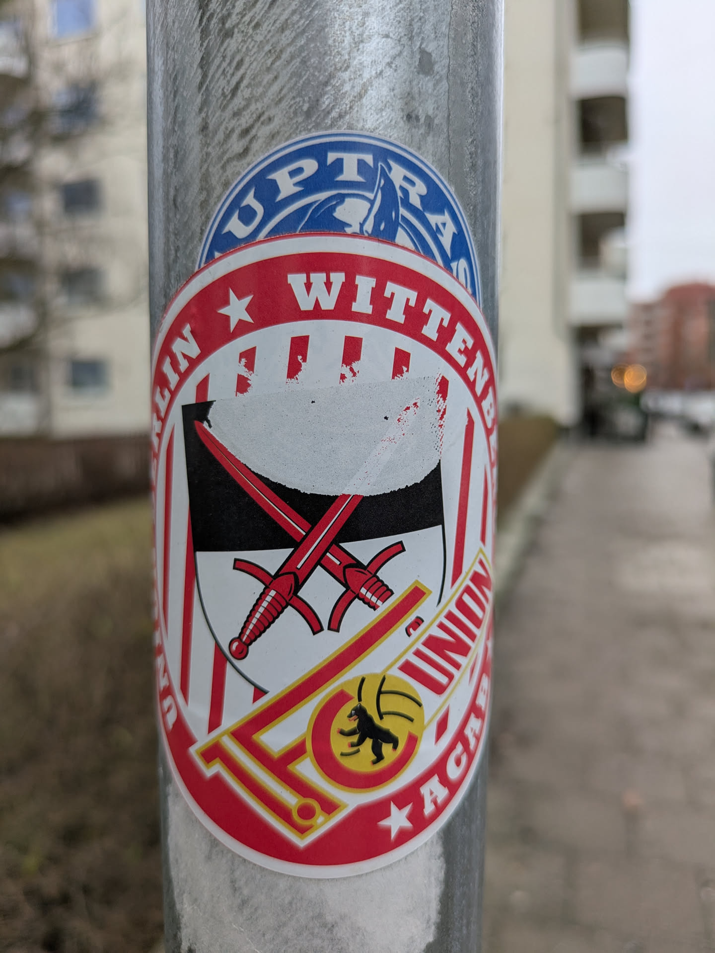 The sticker features the 1. FC Union Berlin academy logo. The logo is round with red and white stripes, and the text "BERLIN WITTENAU" is printed in red, surrounding the coat of arms of Wittenau, Berlin. Two swords are positioned behind the coat of arms. Below, the team's logo is situated within the 'FC' abbreviation. Lastly, the word "ACADEMY" is also written in red.